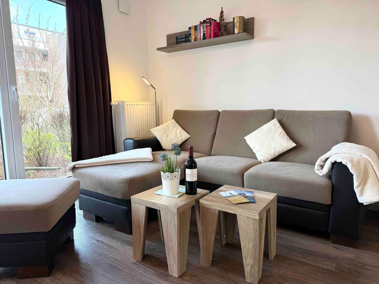 Ein Wohnzimmer mit Sofa, zwei Tischen und einem Fenster. Auf dem Sofa liegen Kissen und eine Decke.