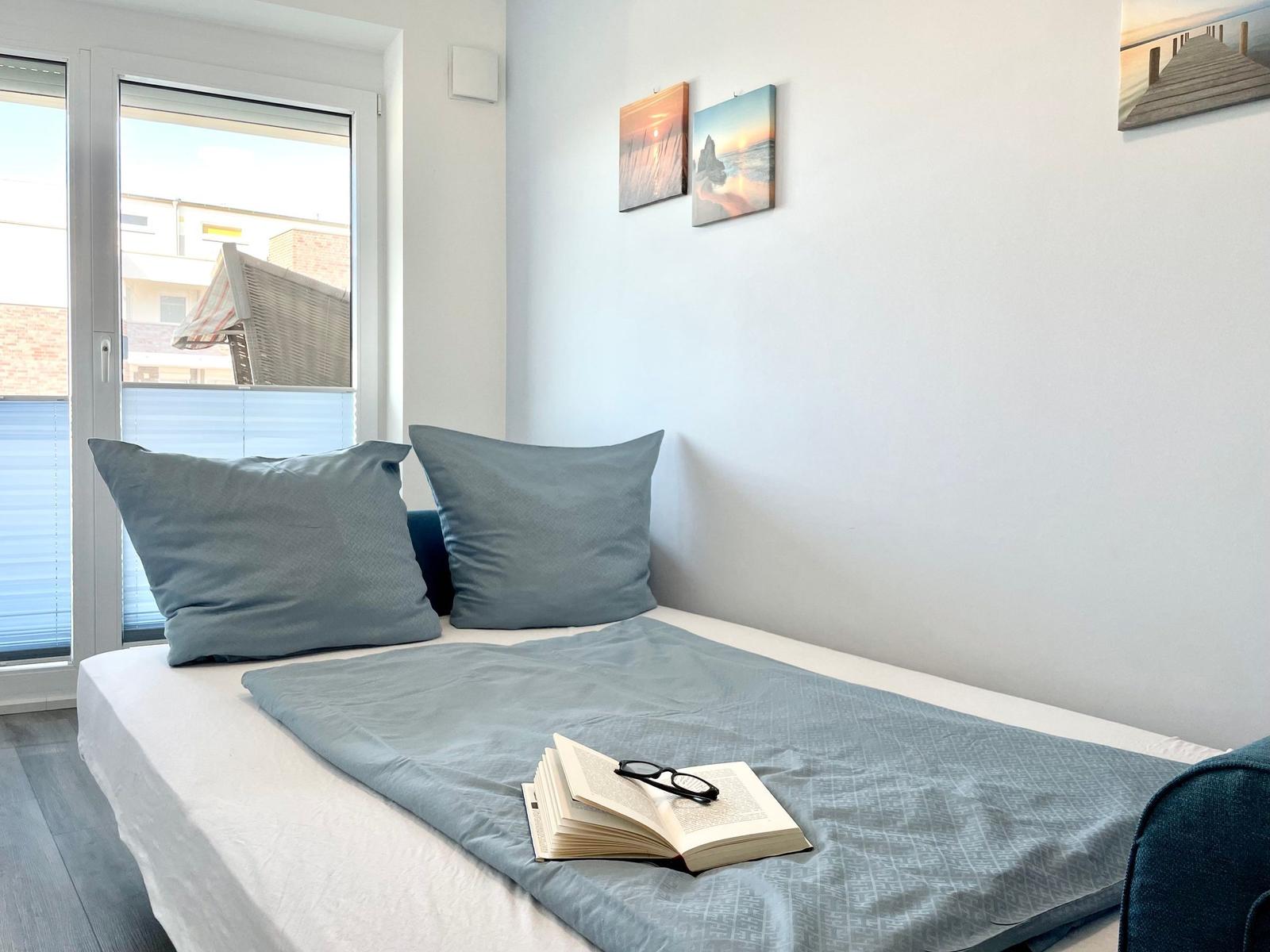 Ein Bett mit grauer Bettwäsche, zwei Kissen und einem Buch mit Brille.