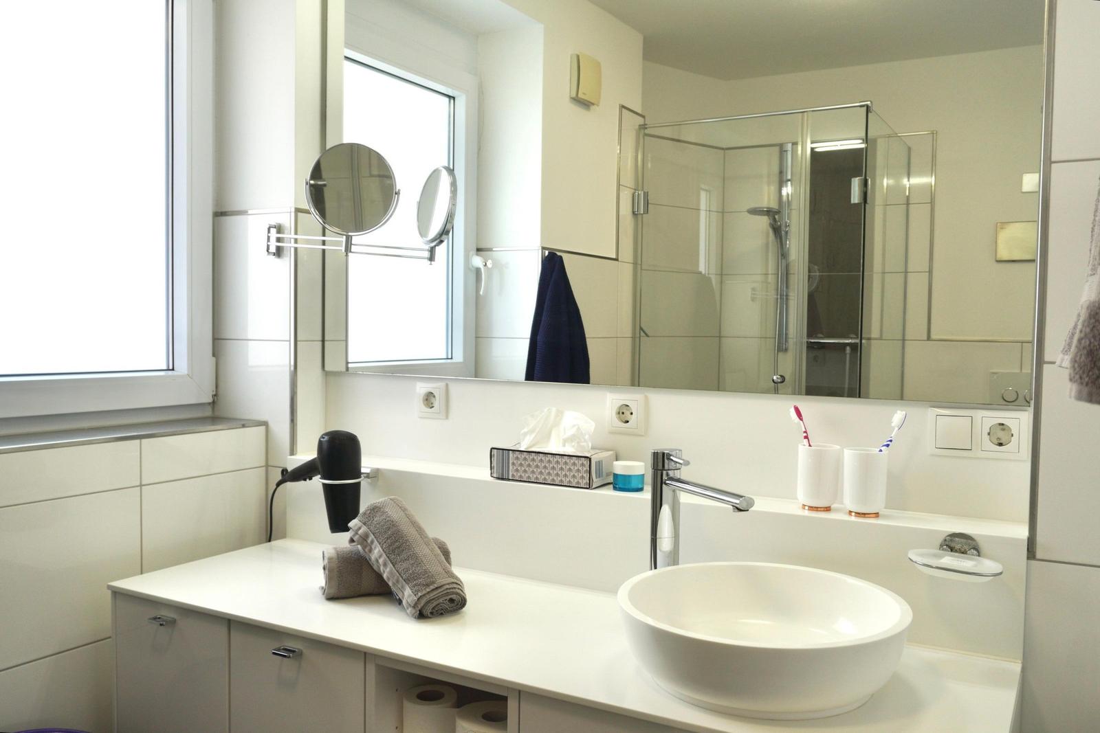 Badezimmer mit Waschtisch, Spiegel und Dusche. Fenster, Handtuch und Haartrockner sichtbar.