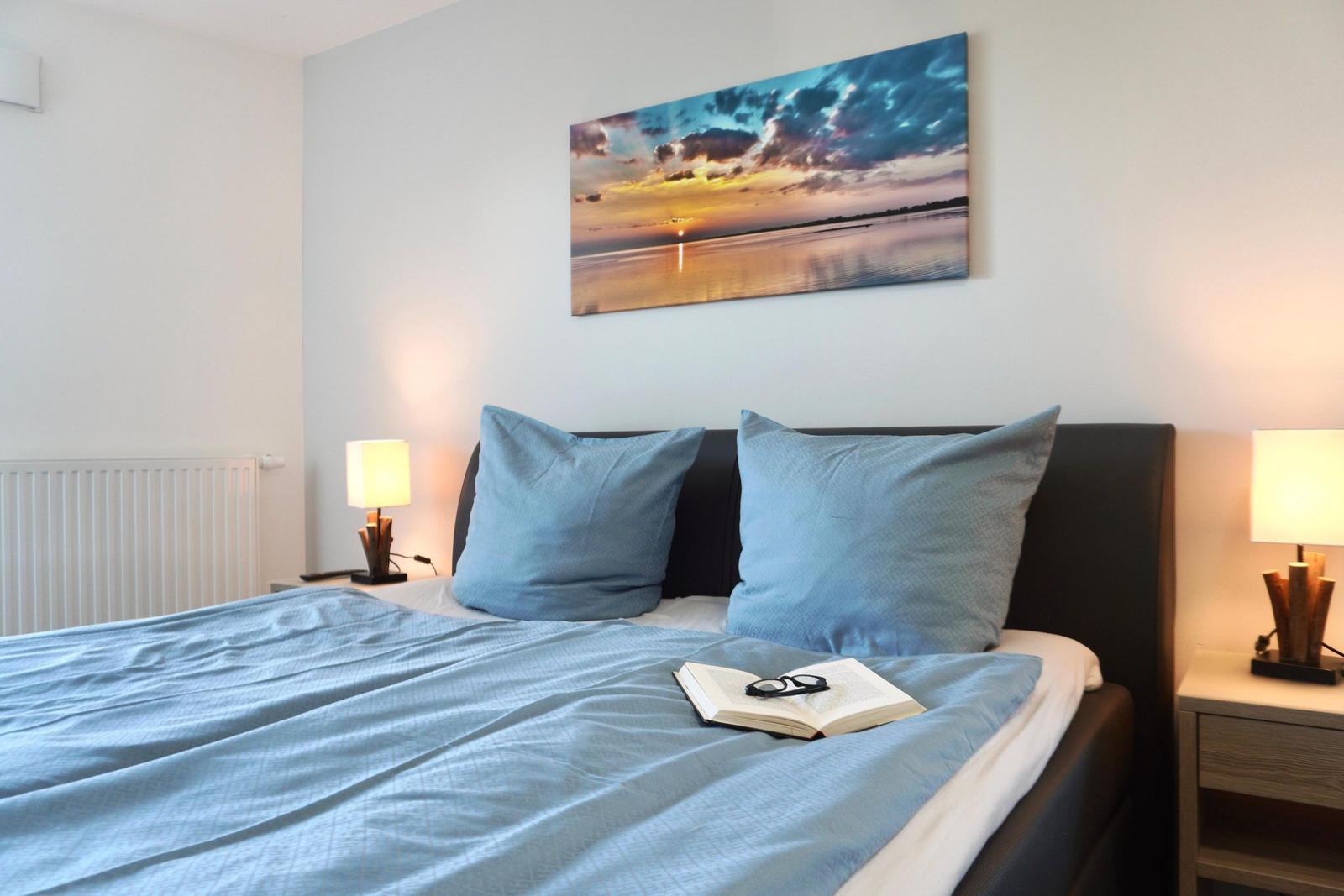 Schlafzimmer mit Bett, zwei Nachttischen und Wandbild. Auf Bett liegt Buch mit Brille.