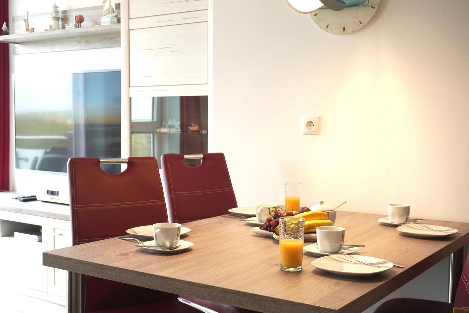 Dining table with four chairs, fruit, and drinks. Wall clock and window in background.