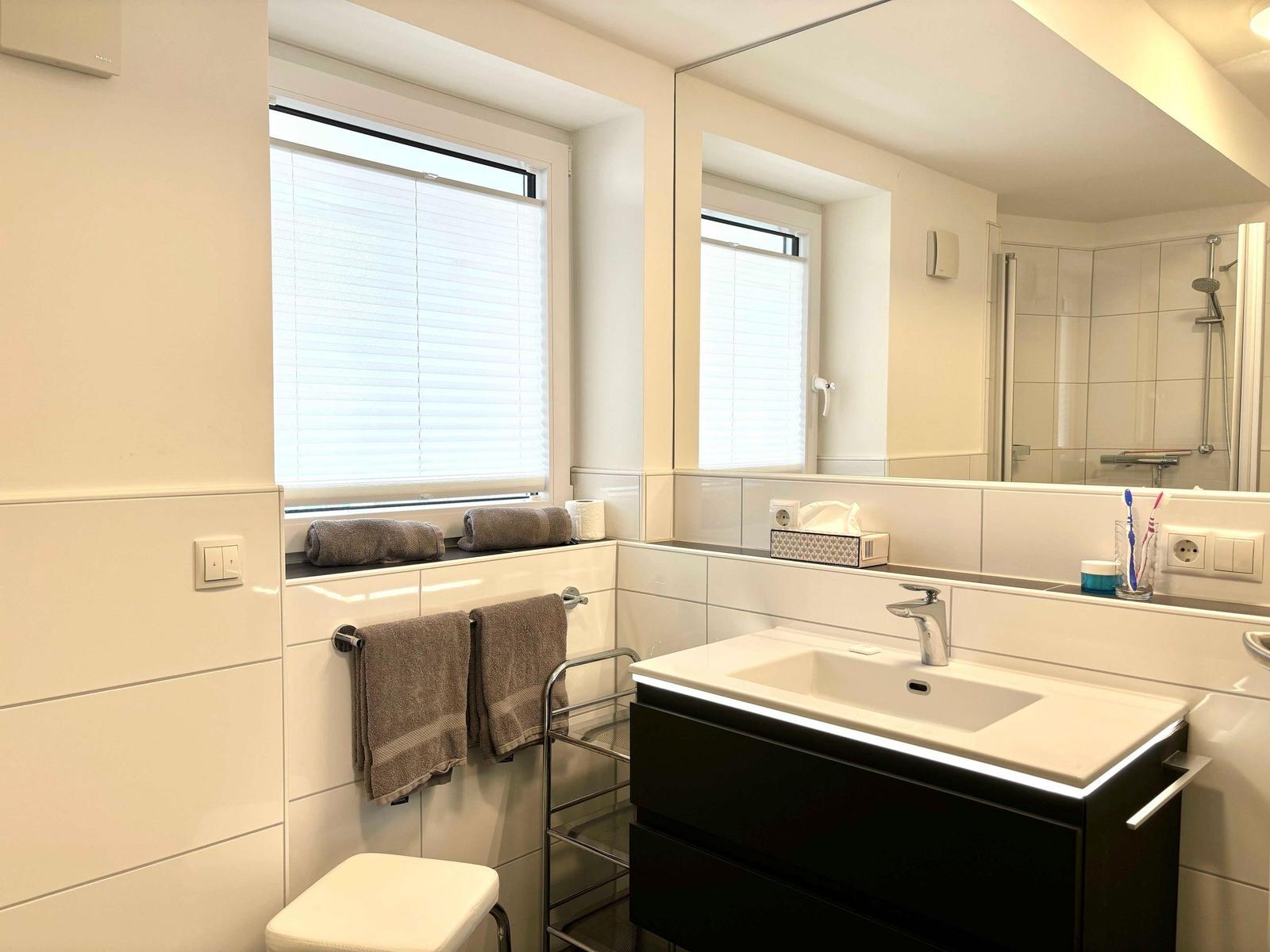 Bathroom with sink, mirror, shower, and window with blinds.
