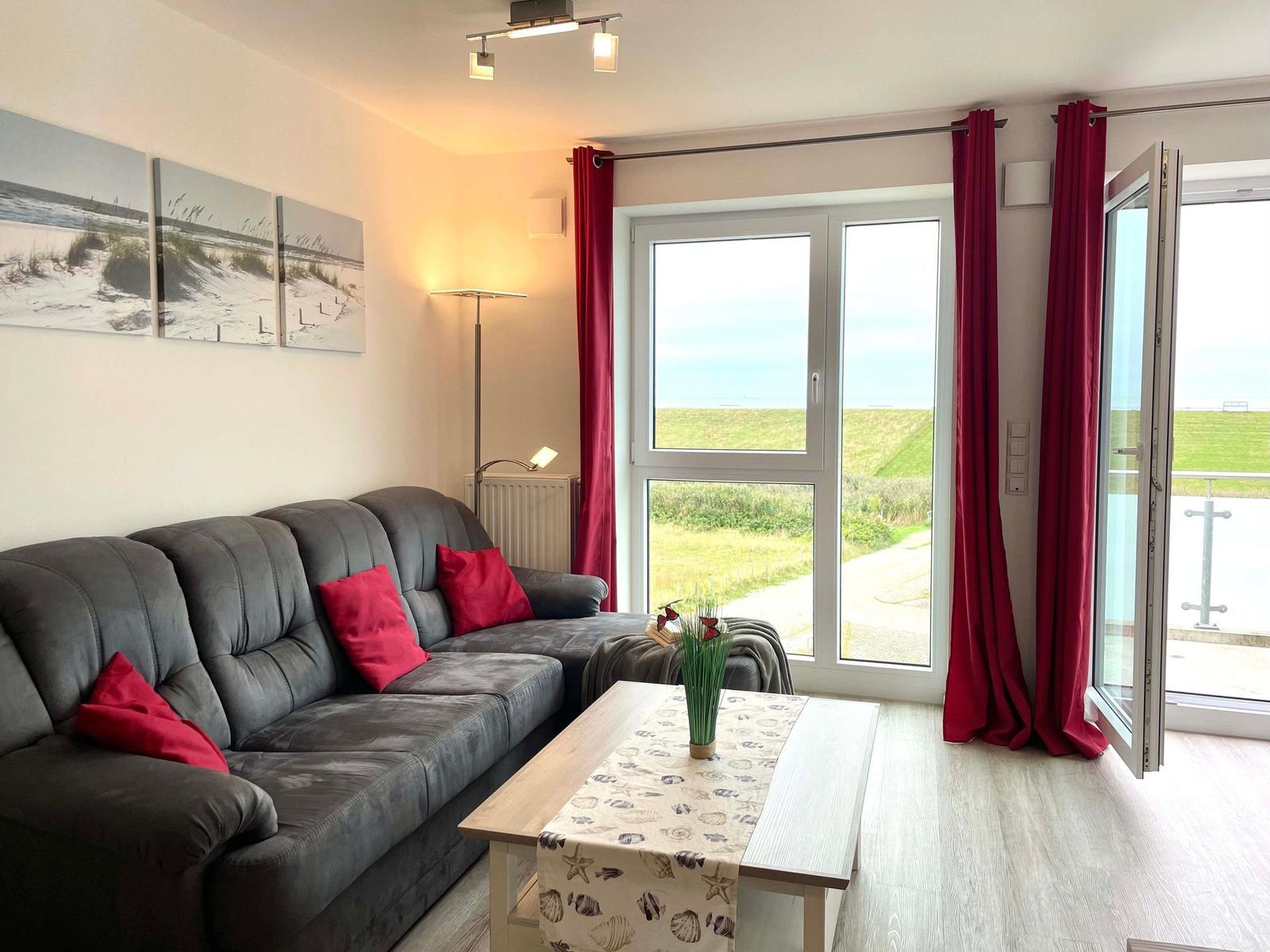 Living room with sofa, table, and view of green field. Red curtains and wall art.