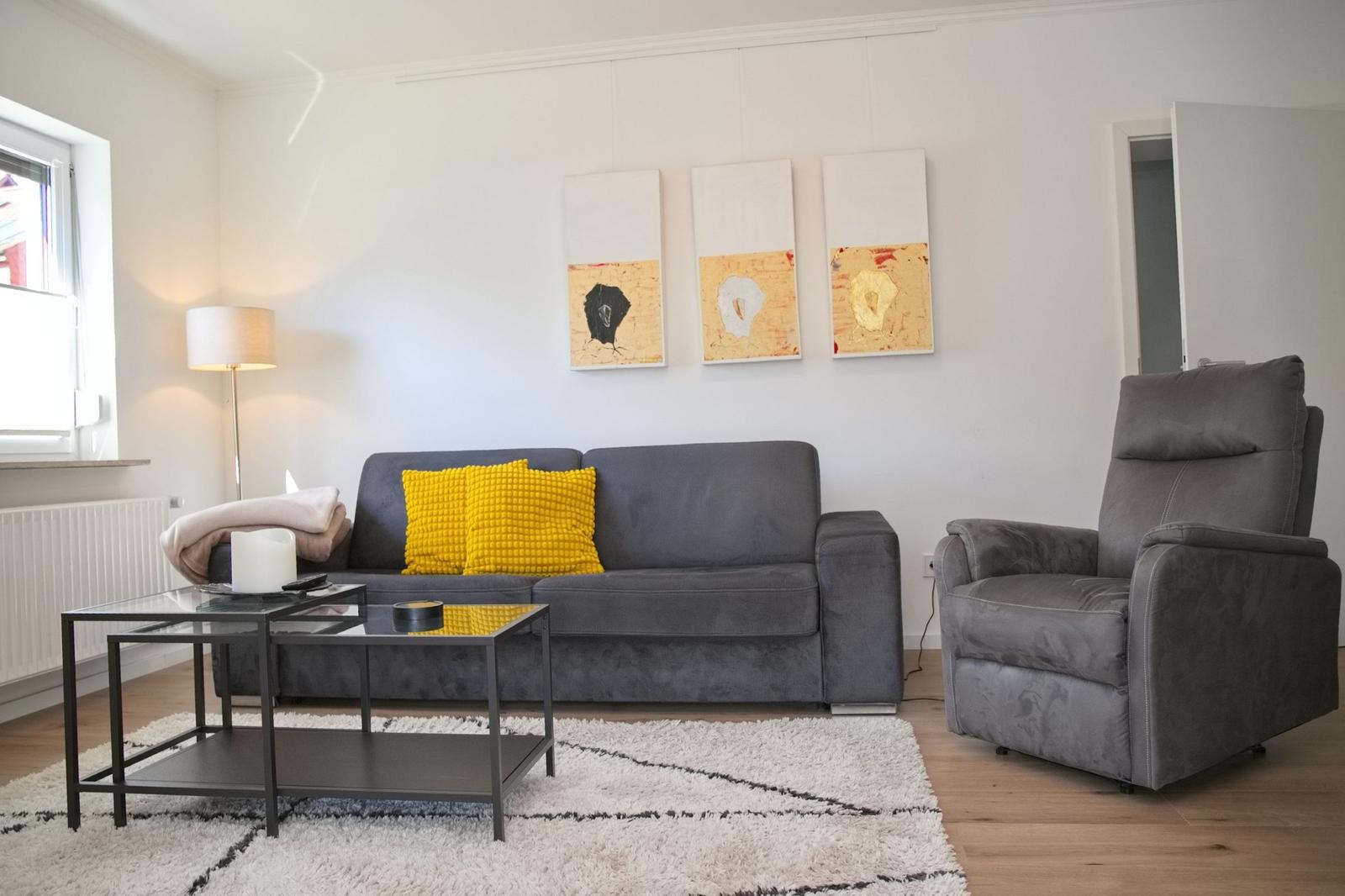 Ein Wohnzimmer mit grauem Sofa, gelben Kissen und einem Sessel. An der Wand hängen drei Bilder.