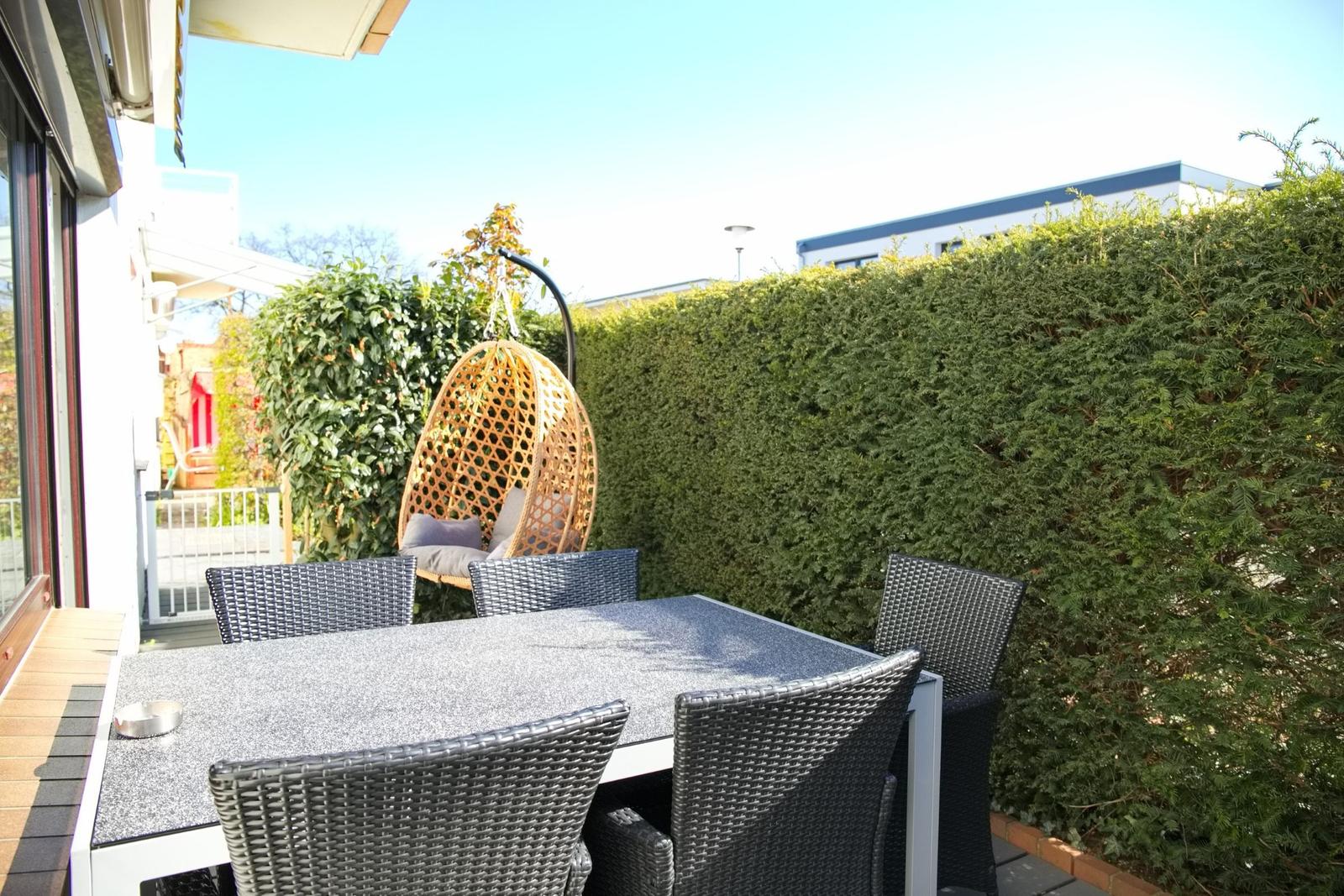 Terrasse mit Esstisch, Stühlen und Hängesessel hinter Hecke.
