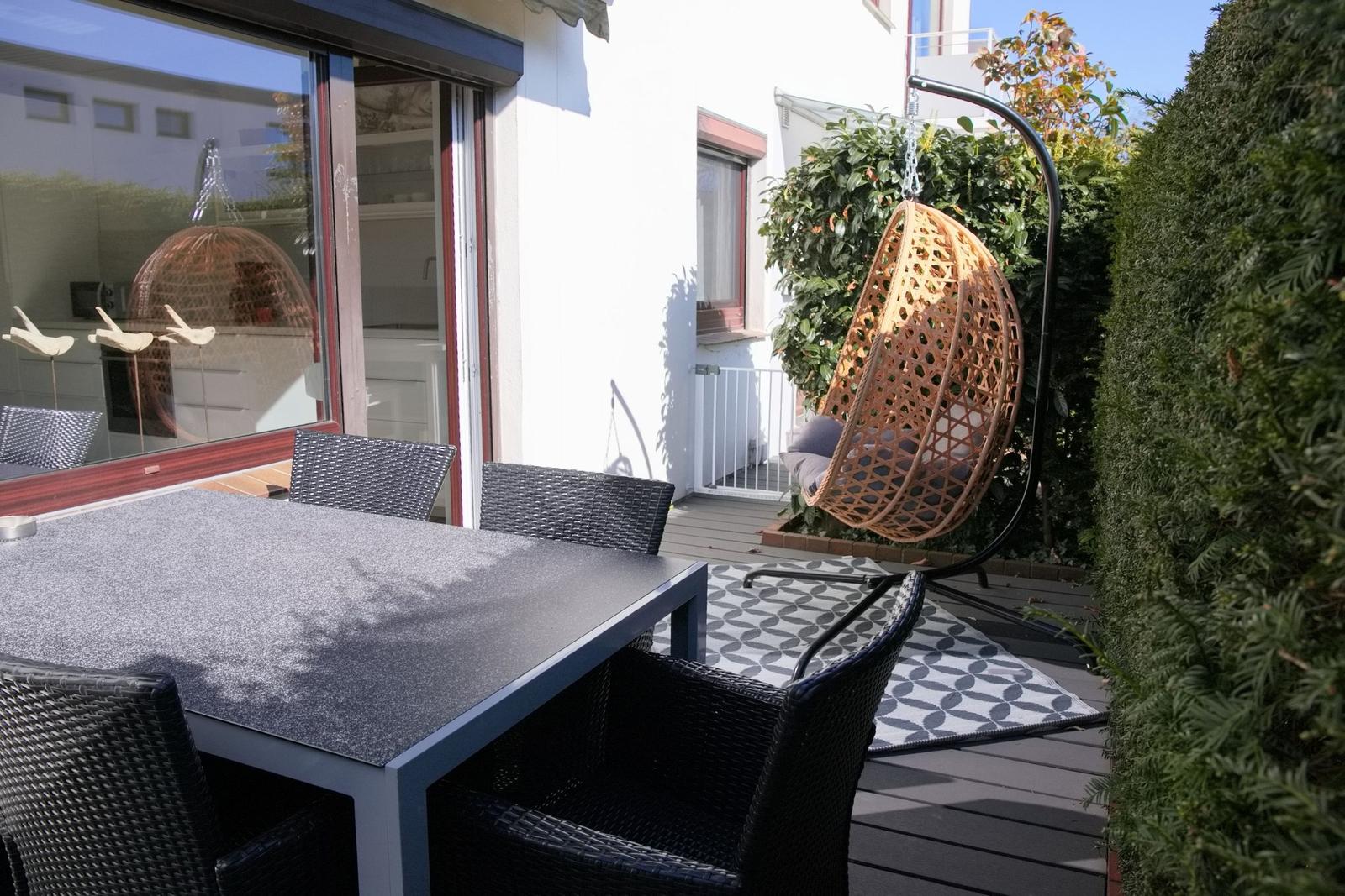 Terrasse mit Tisch, Stühlen und Hängesessel. Durchgehendes Glasfenster zur Küche.