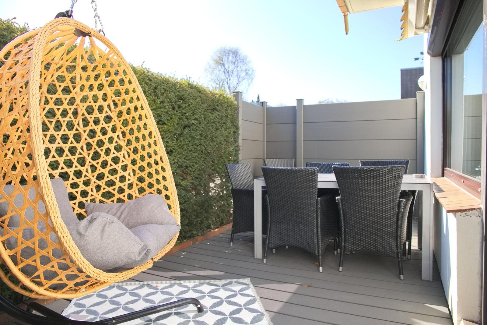 Terrasse mit Hängesessel, Esstisch und Stühlen. Hintergrund: Hecke und Zaun.