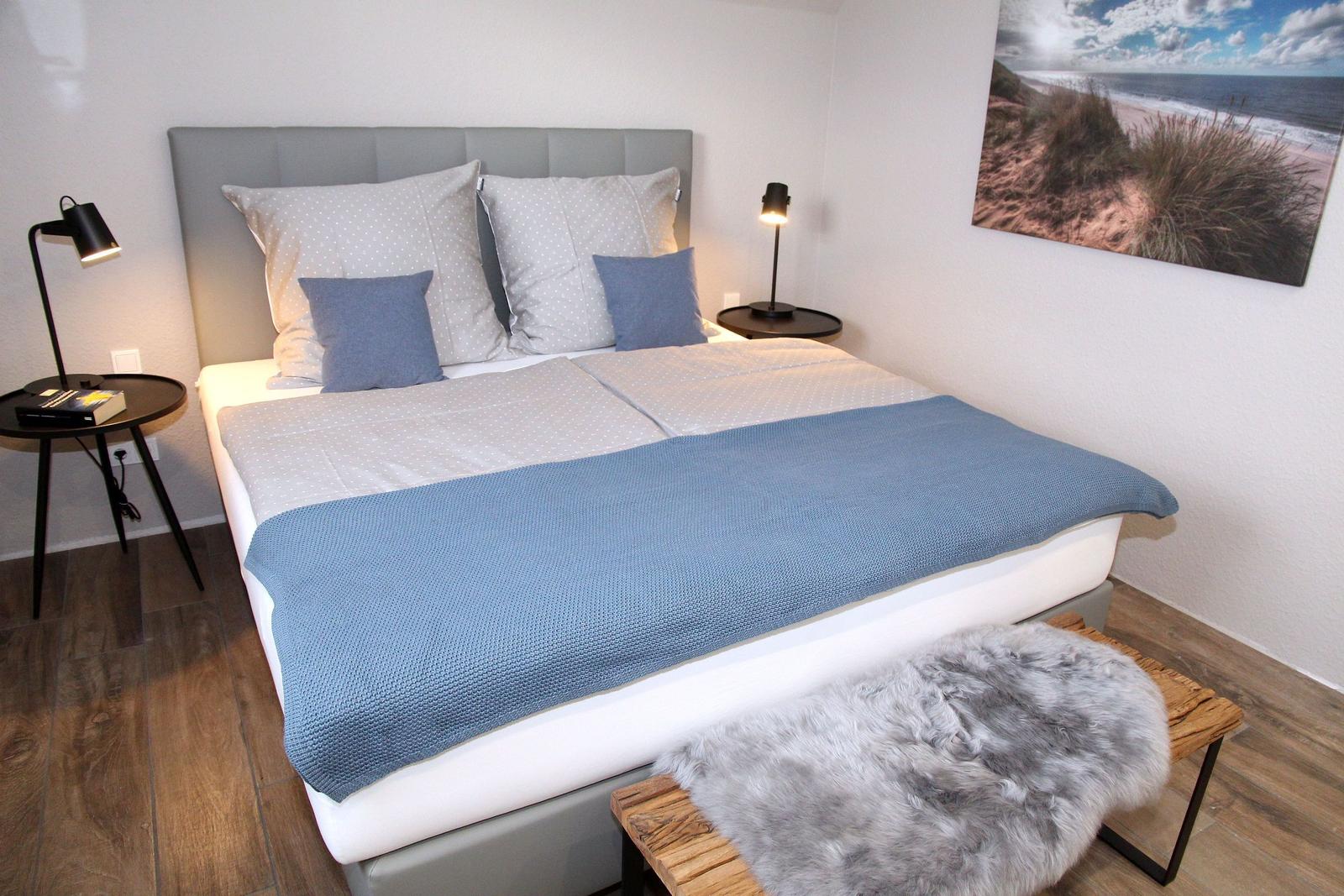 Doppelbett mit blauer Decke, zwei Nachttische und Wandbild mit Strandmotiv.