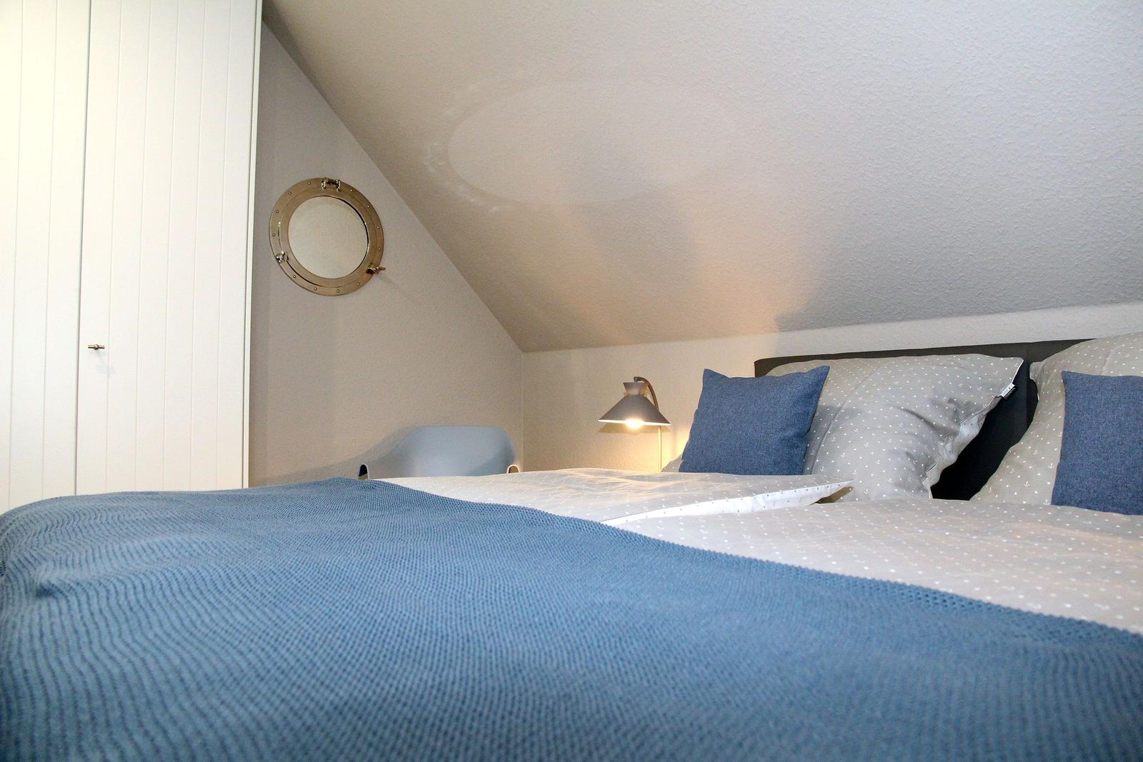 Schlafzimmer mit Bett, blauer Decke und runder Fensteröffnung an schräger Decke.
