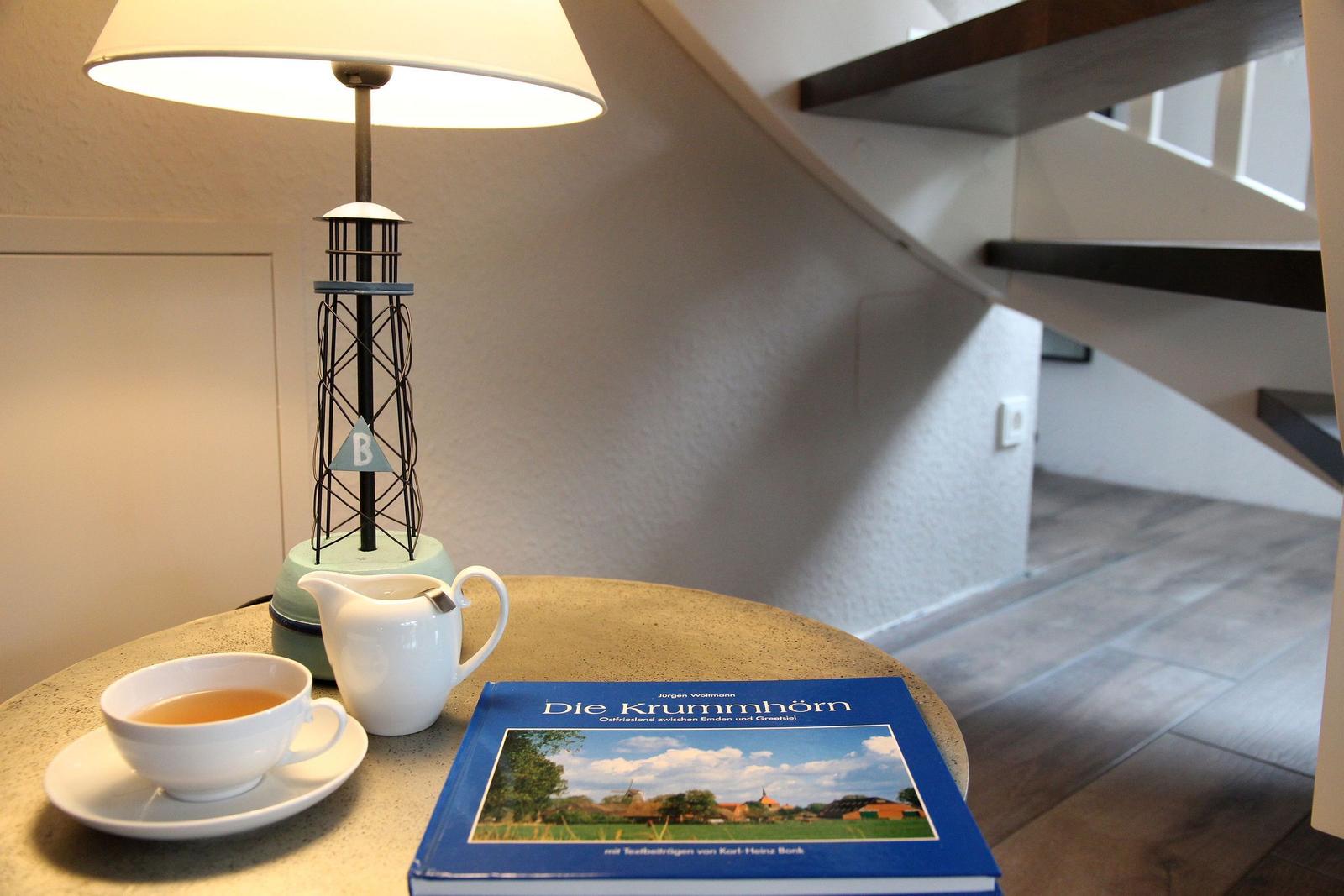 Tisch mit Tasse, Milchkanne und Buch unter einer Lampe neben einer Treppe.