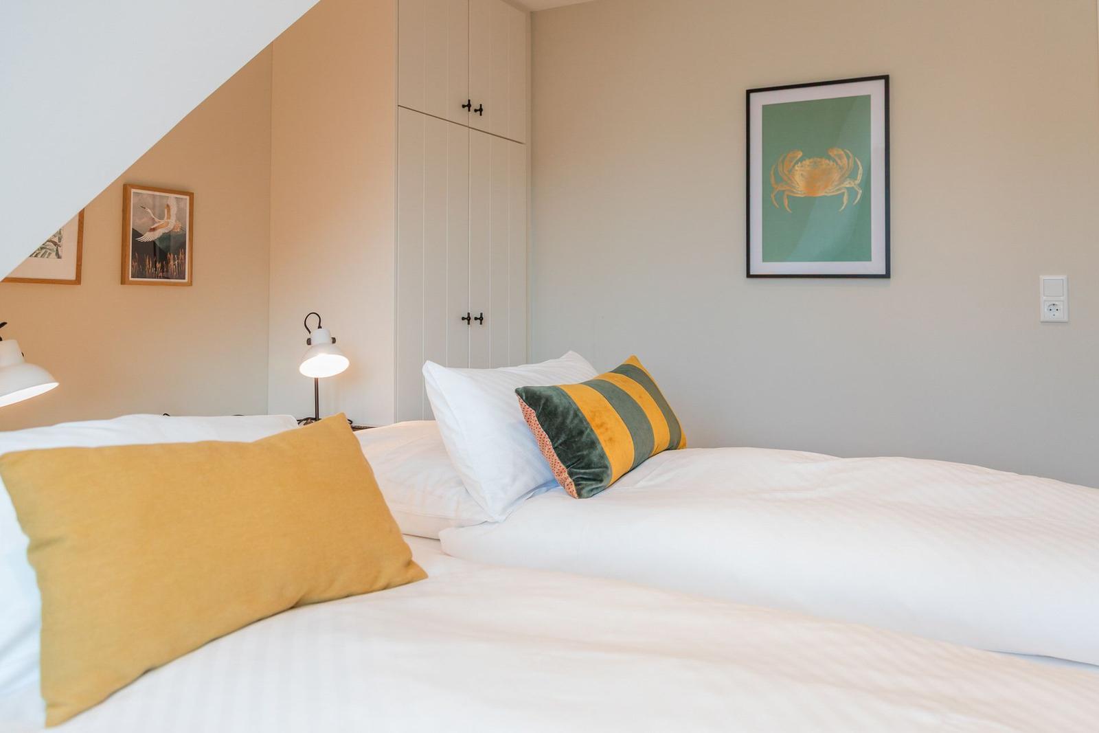 Schlafzimmer mit Bett, Kissen und Wandbildern. Hinter dem Bett steht ein Schrank.