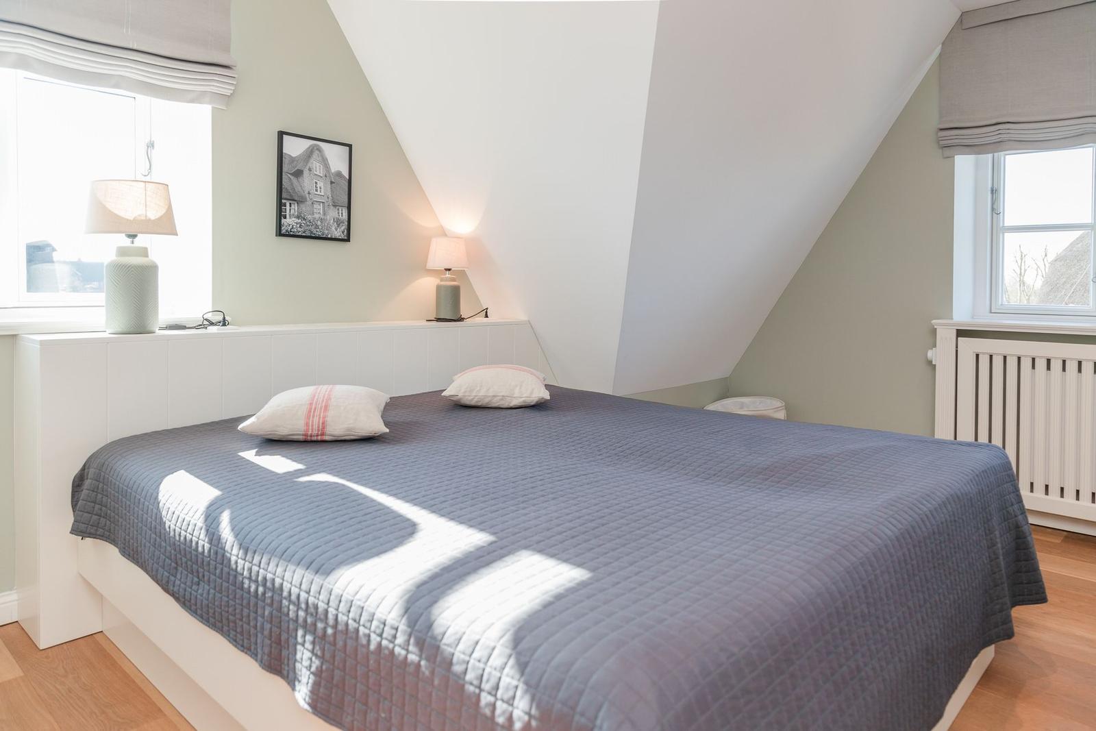 Schlafzimmer mit Dachschräge, Bett, zwei Fenster und Lampen.