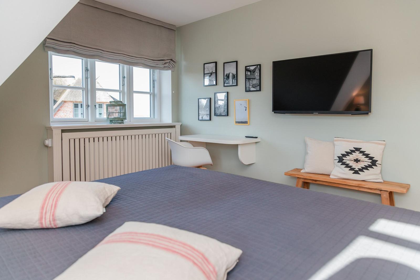 Schlafzimmer mit Bett, TV, Schreibtisch und Fenster mit Blick auf Dächer.