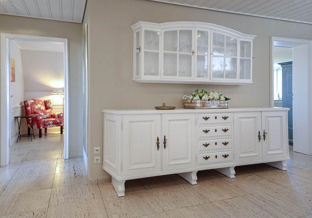 Weißer Sideboard mit Schubladen und darüber hängendem Schrank. Im Hintergrund ist ein Wohnzimmer mit Sessel sichtbar.