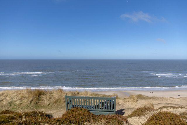 Ein grüner Holzstuhl steht vor Dünen und blickt auf das Meer.