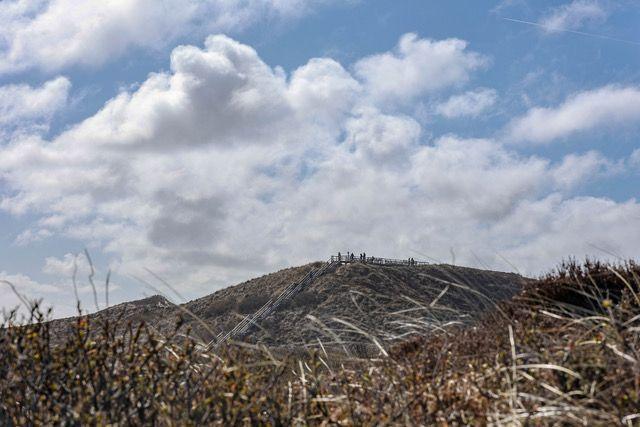 Blick auf einen Berg mit Wanderweg und Personen am Gipfel unter bewölktem Himmel.