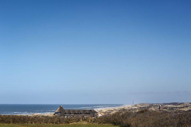 Haus mit Dach nahe Strand und Dünen unter blauem Himmel.