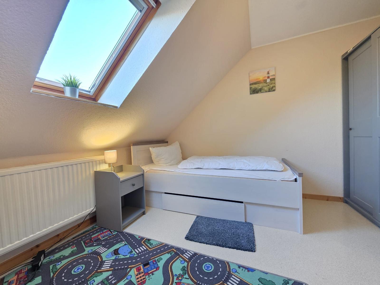 Schlafzimmer mit Dachschräge, Dachfenster, Bett, Nachttisch und Kinderbildteppich.
