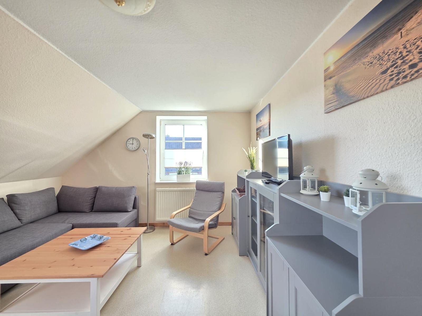 Wohnzimmer mit Sofa, Sessel, TV und Schrank. Dachschräge und Fenster sichtbar.