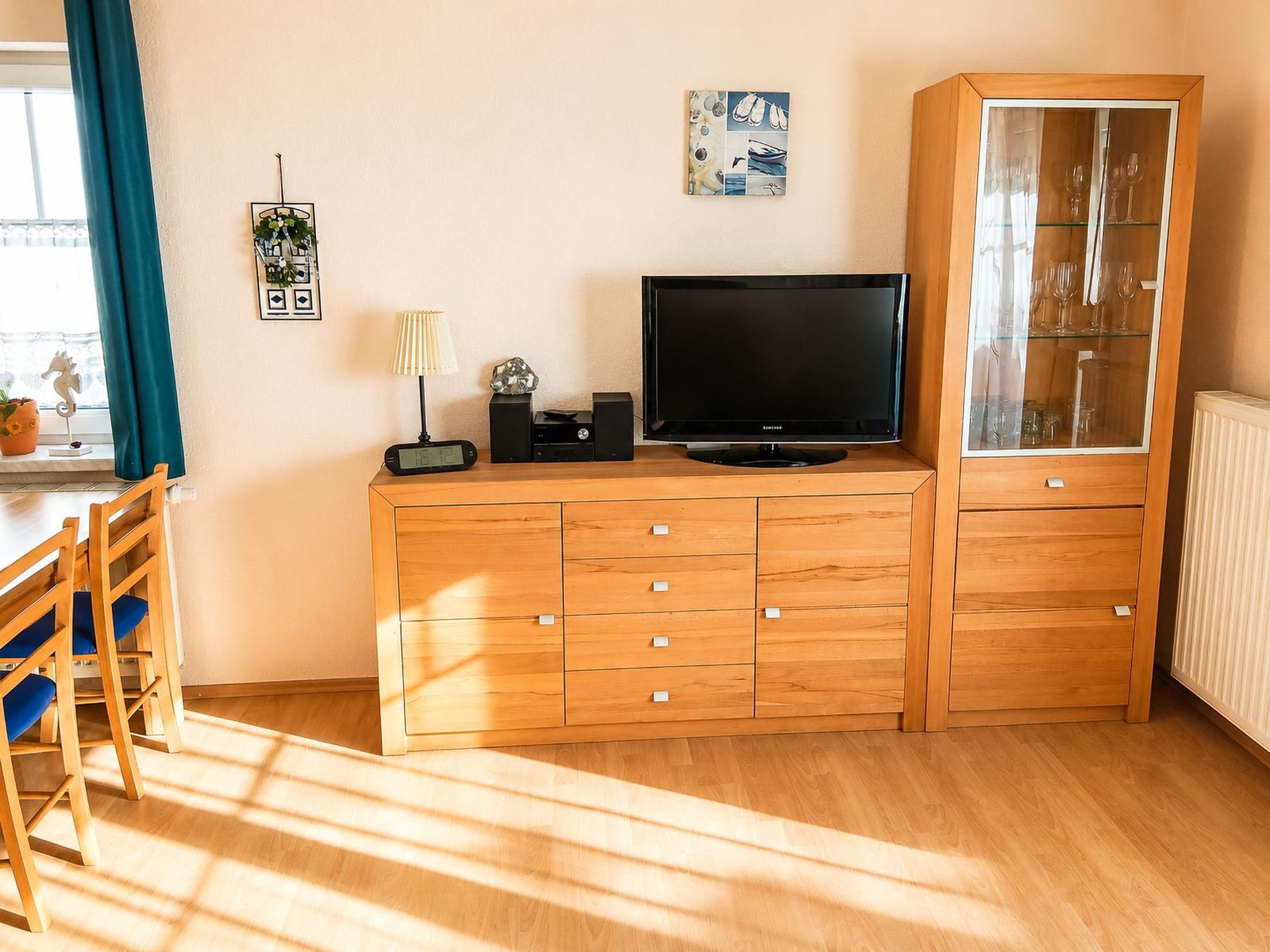 Ein Wohnzimmer mit Holzmöbeln, Fernseher und Glasvitrine. Fenster mit blauen Vorhängen.