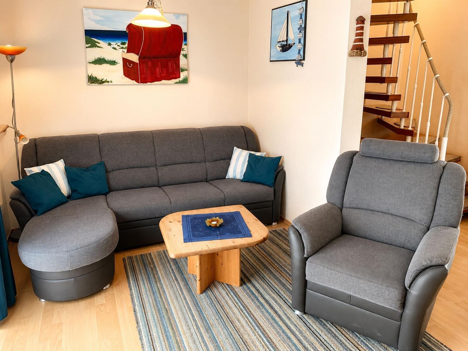 Wohnzimmer mit grauem Sofa, Sessel und Holztisch. Wandbilder und Treppe sind sichtbar.