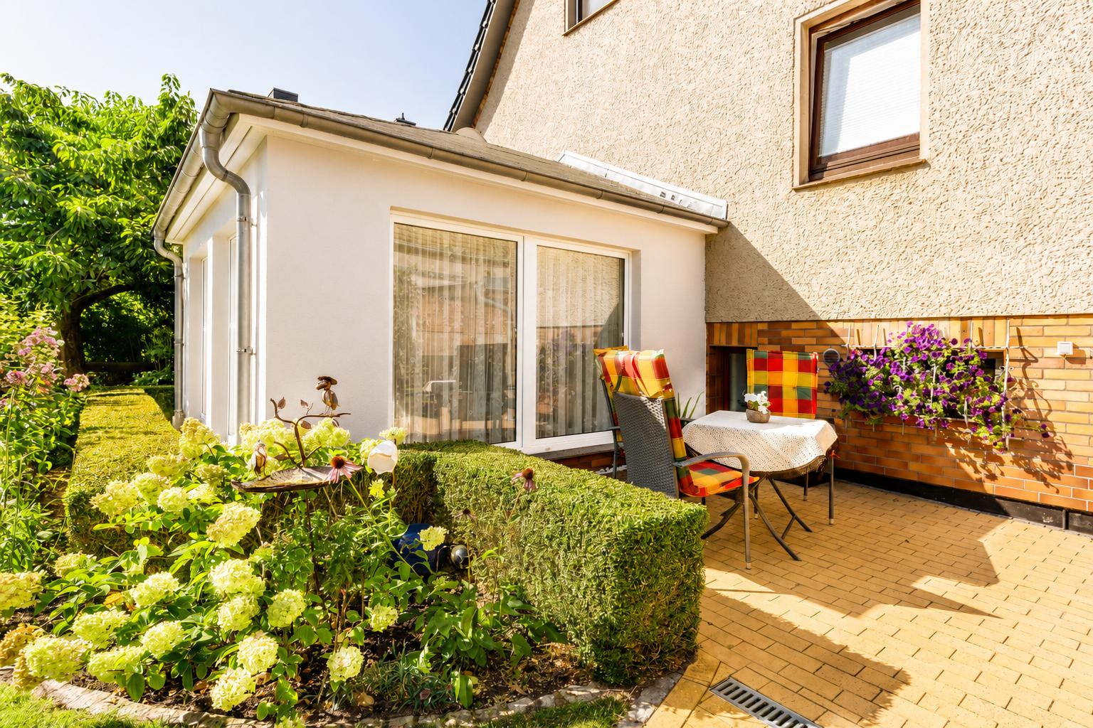 Terrasse mit Tisch, Stühlen und Blumenbeeten. Hintergrund: Haus mit Fenstern.