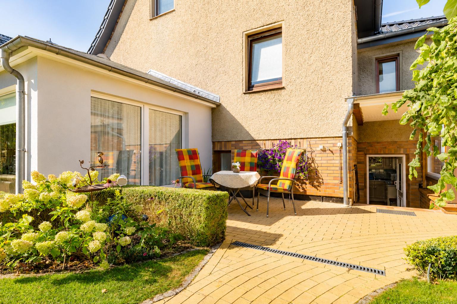 Terrasse mit Tisch, drei Stühlen und Blumenbeete. Hinterhof mit Gartentüre und Fenster.