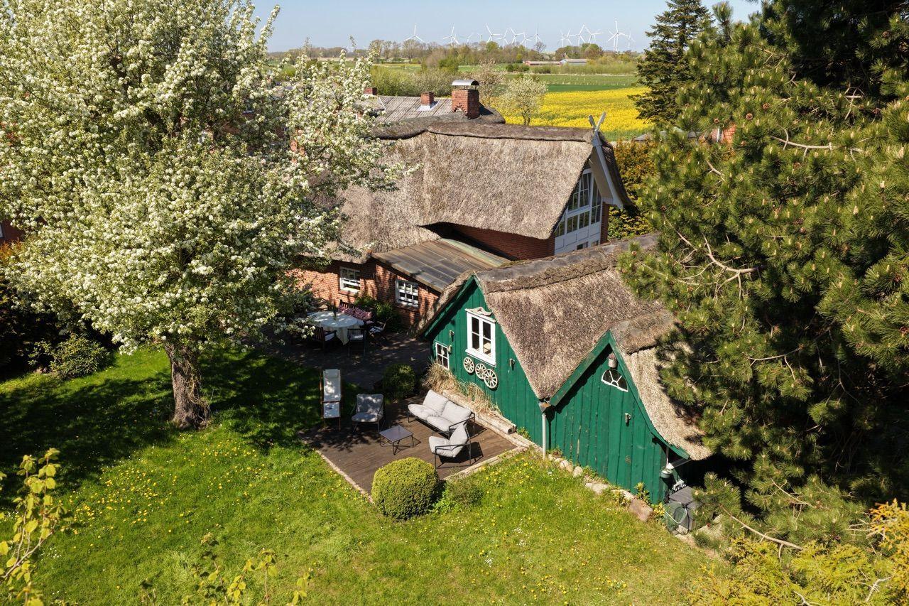 Dorfhaus mit strohgedecktem Dach, Garten und Terrasse. Im Hintergrund Windräder und Felder.