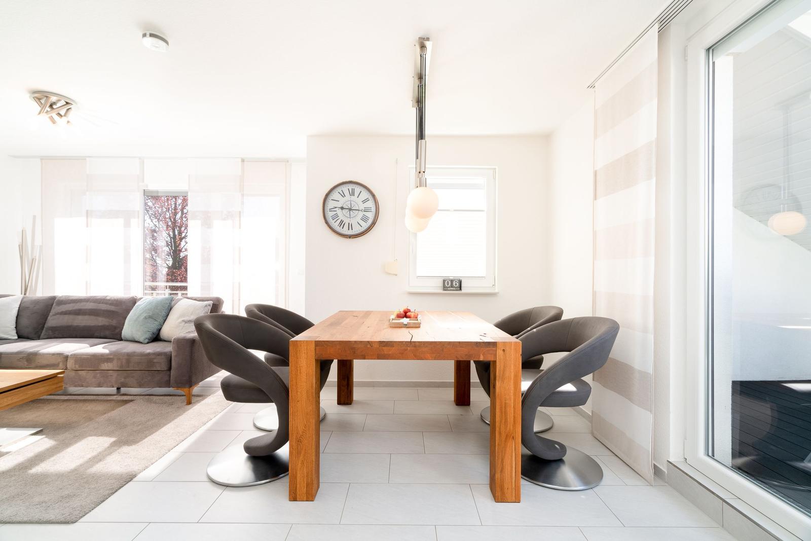 Essbereich mit Holztisch und vier Stühlen. Im Hintergrund Wohnbereich mit Sofa und Fenster.
