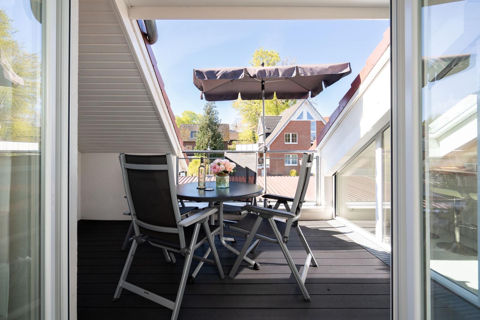 Terrasse mit Tisch, zwei Stühlen und Sonnenschirm. Blick auf Wohnhaus und Bäume.