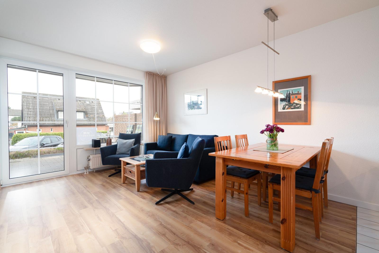 Living and dining area with wooden floor, blue sofa, wooden table and chairs. Large window front with view outside.