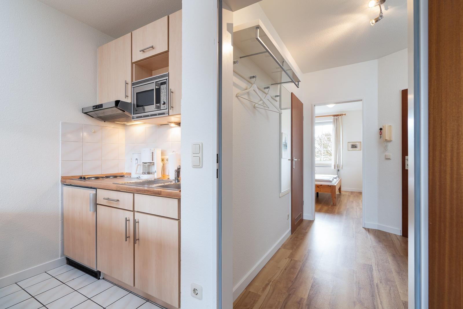 Kitchen with microwave and sink. Hallway with coat hooks and bedroom in the background.