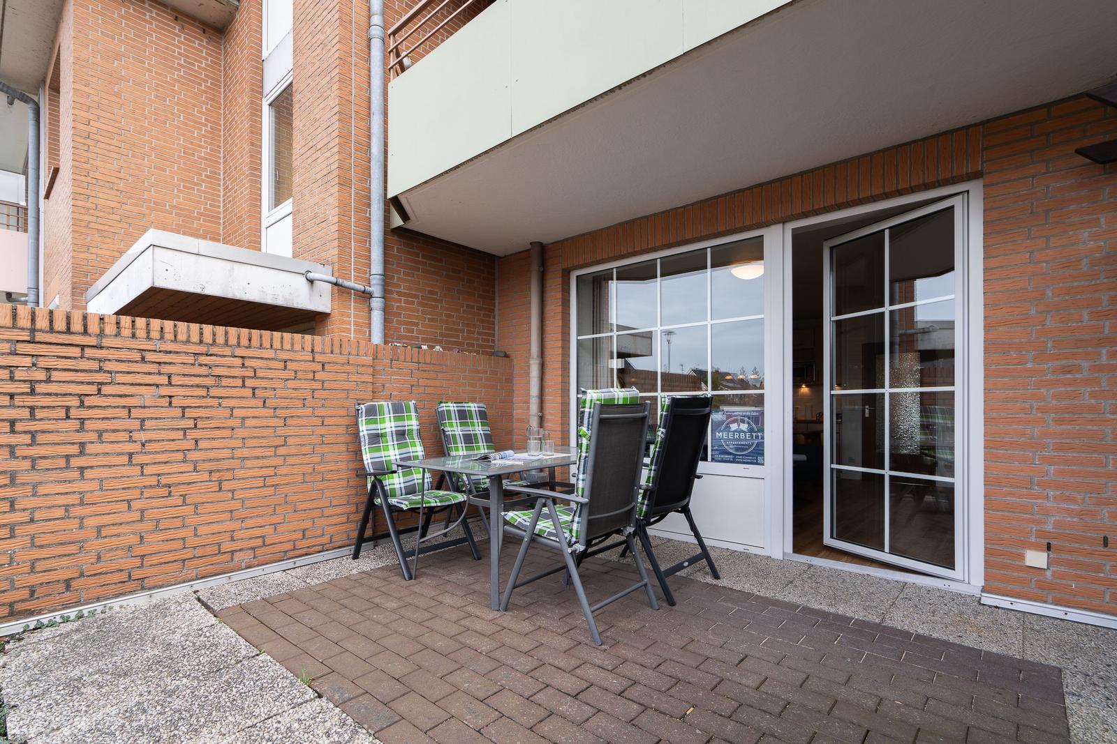 Terrace with table and chairs before a glass door. Brick wall and paved floor.