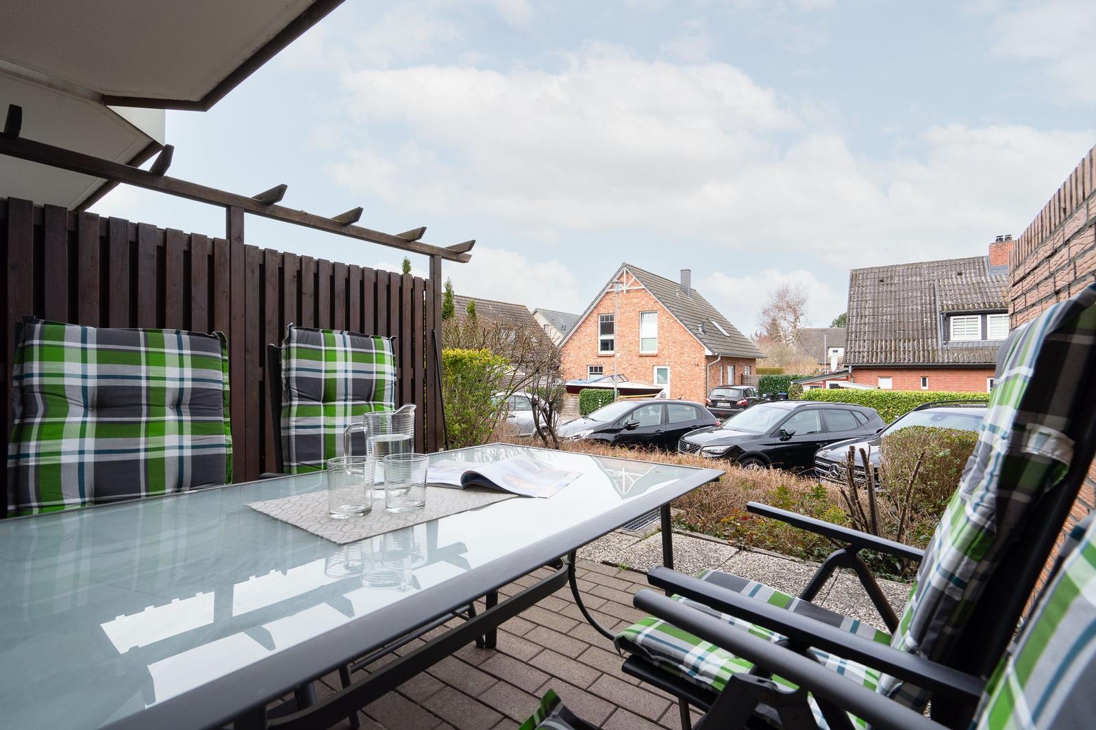 Terrace with glass table and green-white cushions. Houses and parked cars are visible in the background.