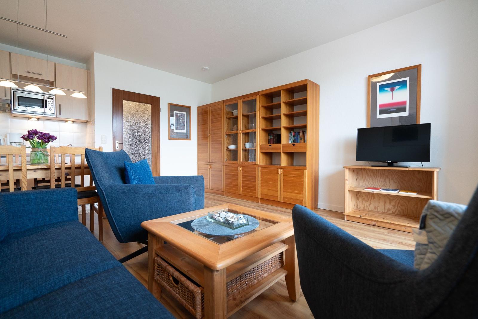 Living room with blue chairs, wooden furniture, and TV. Kitchen in background.