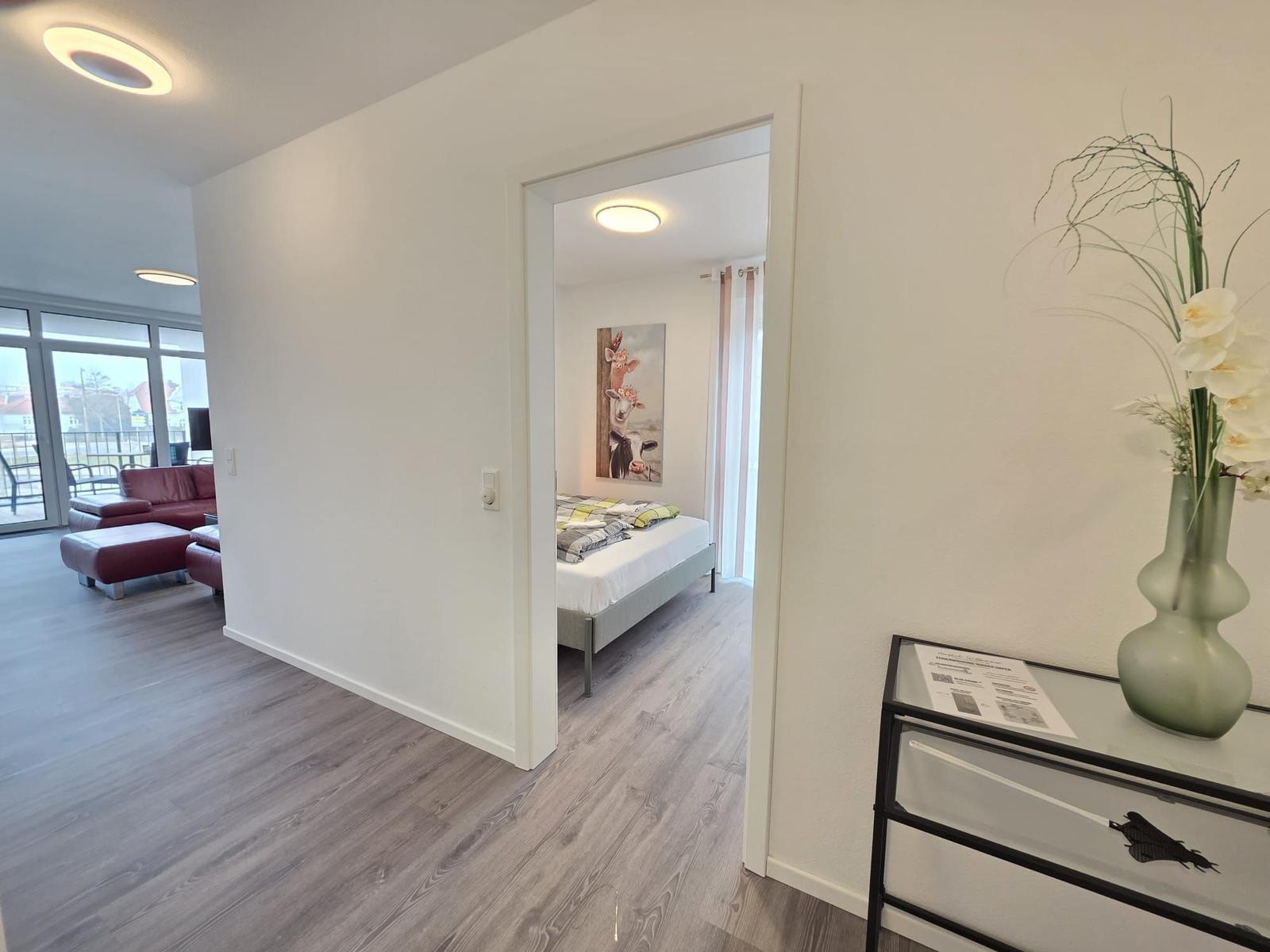 Ein Schlafzimmer mit Bett und Wandbild ist über einen Flur erreichbar. Im Hintergrund liegt ein Wohnbereich mit Sofa.