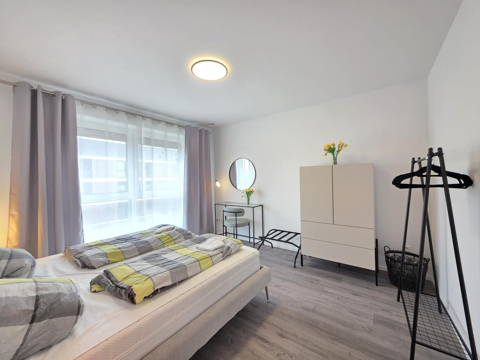 Schlafzimmer mit Bett, Schrank, Kleiderständer und Fenster mit Vorhängen.