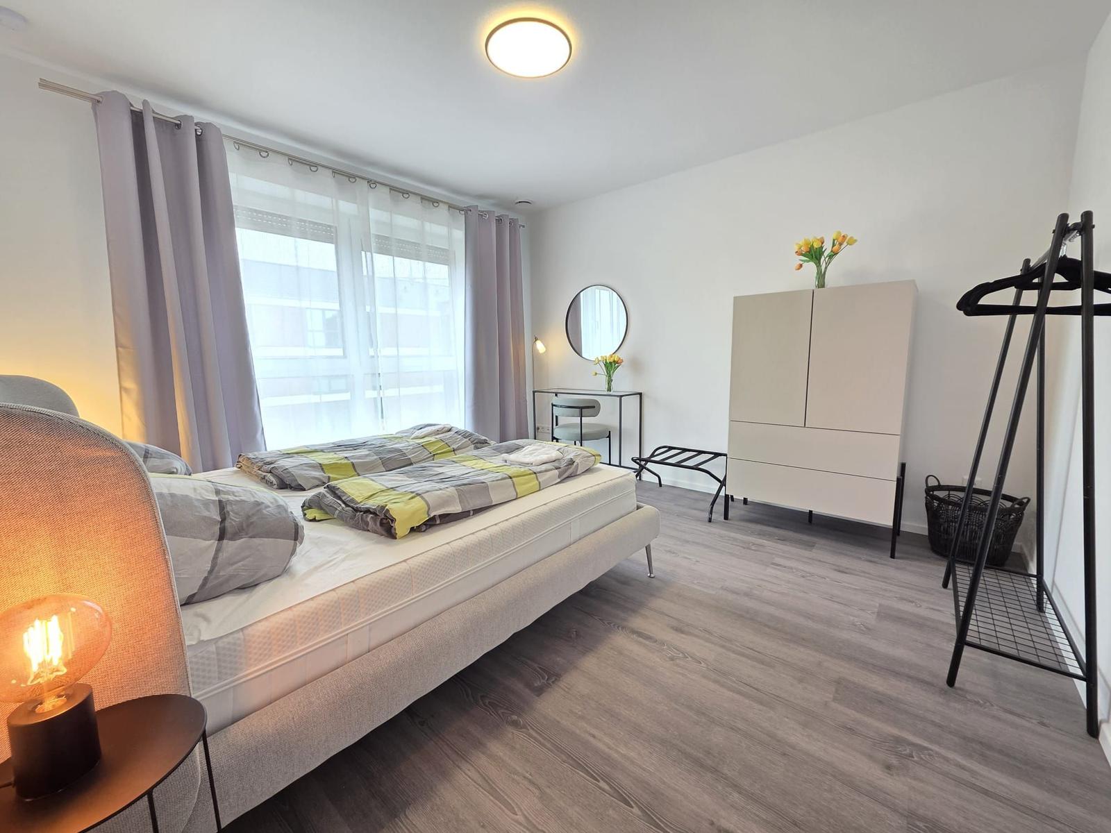 Schlafzimmer mit Bett, Kleiderschrank und Schminktisch. Fenster mit Vorhängen und Bodenbelag.