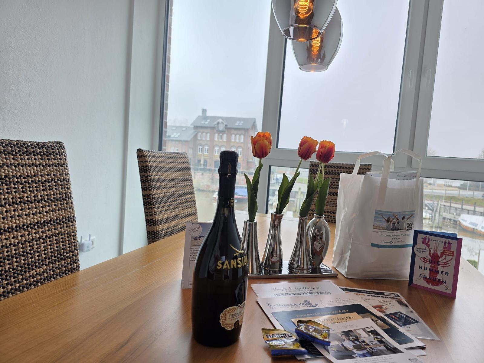 Tisch mit Blumen, Flasche und Informationsmaterial vor großem Fenster mit Aussicht.