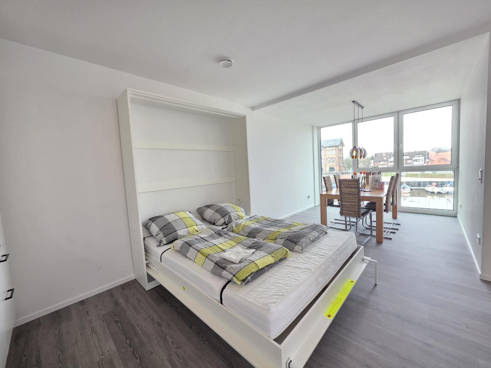 Schlafzimmer mit Schlafwand und Essbereich mit Tisch und Stühlen. Große Fenster mit Blick auf Wasser.