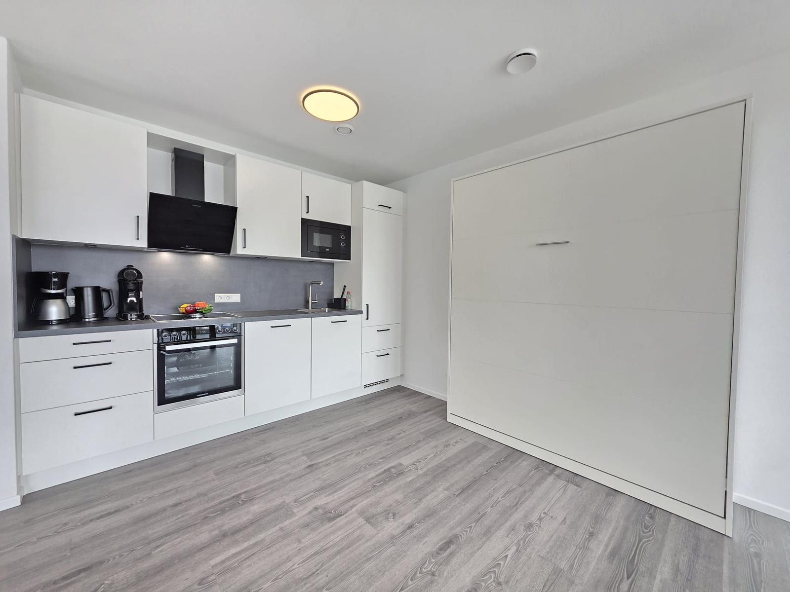Moderne Küche mit weißen Schränken, Herd und Dunstabzug. Holzoptikboden und Schlafwand.