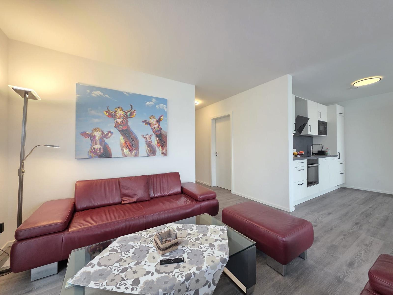 Ein Wohnzimmer mit rotem Sofa, Glas-Tisch und Küchenbereich im Hintergrund.