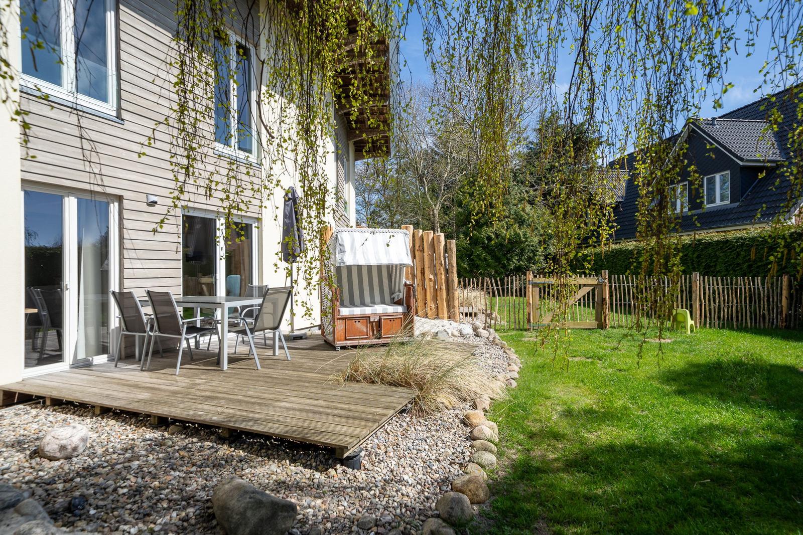 Terrasse mit Tisch, Stühlen und Strandkorb. Grasfläche und Holzzaun im Hintergrund.