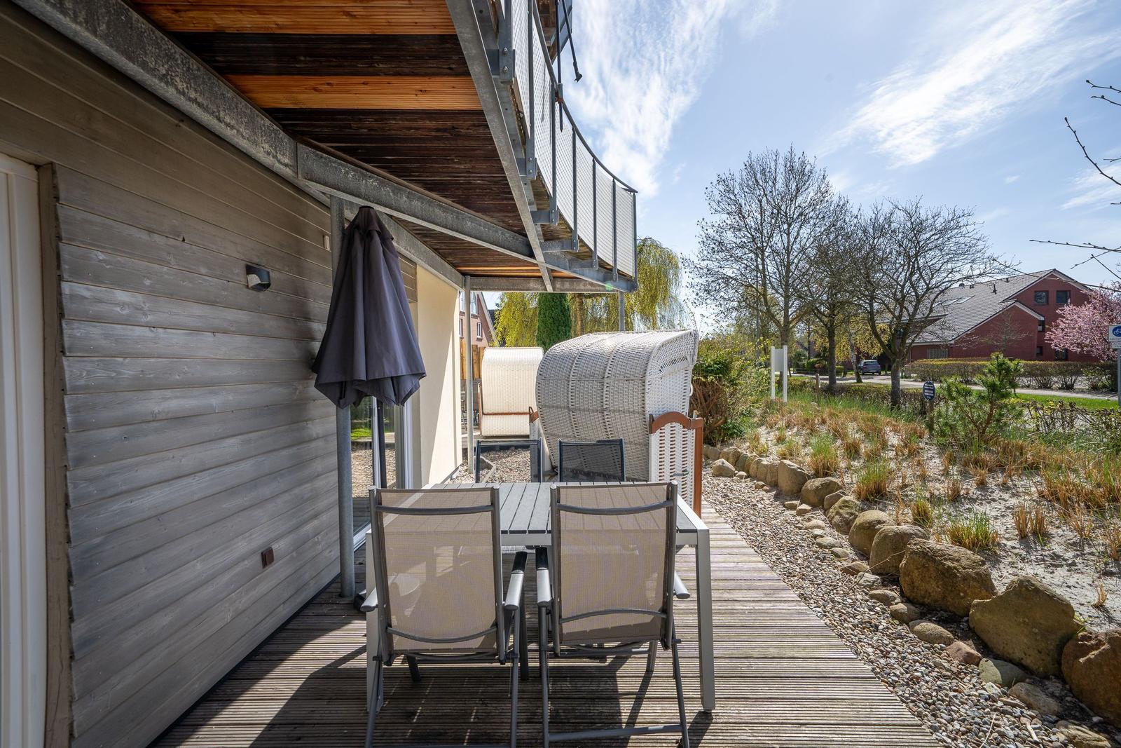 Terrasse mit Tisch, Stühlen und Sonnenschirm neben Holzhaus. Garten mit Steinen und Bäumen.