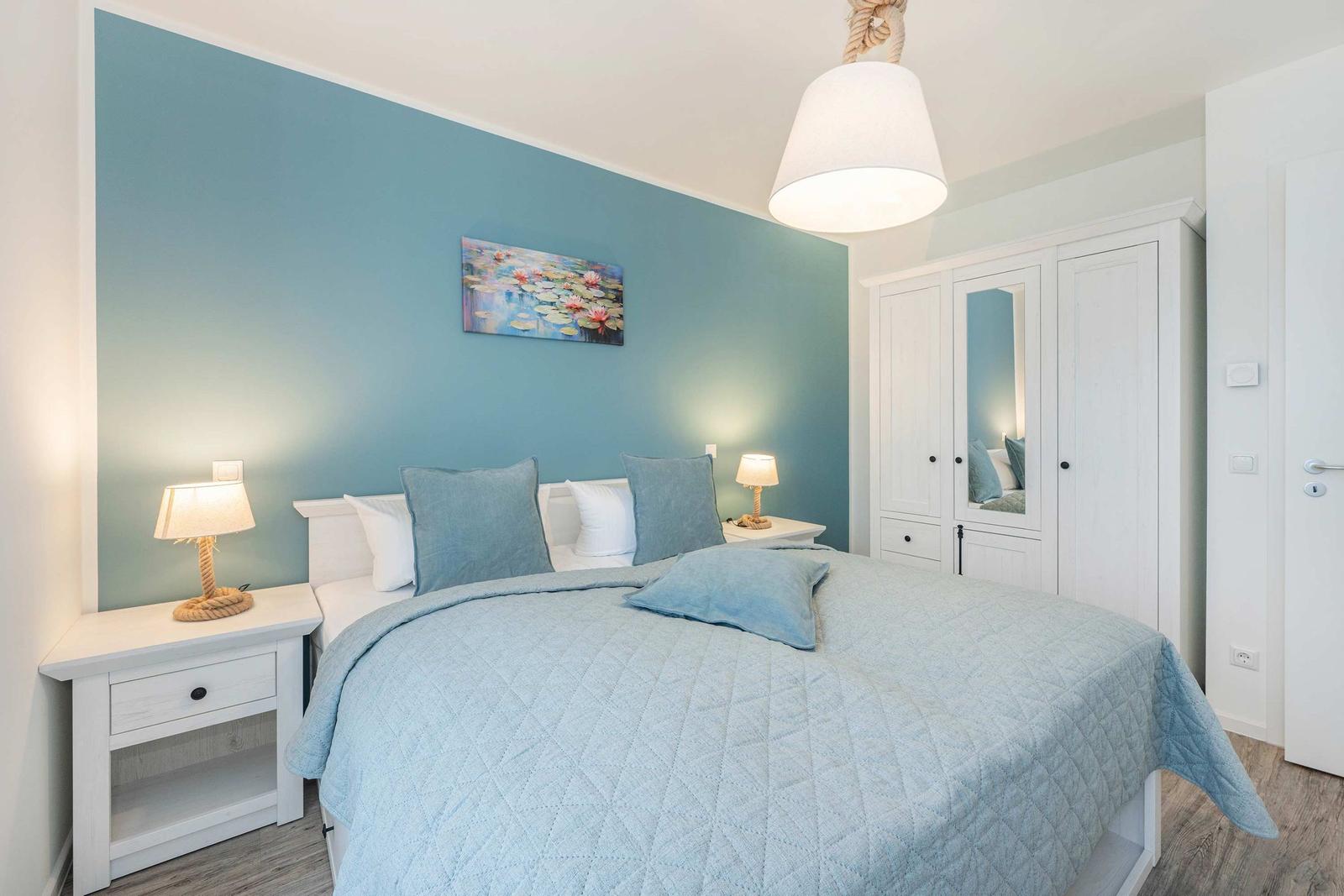 Schlafzimmer mit Bett, Nachttischen und Kleiderschrank. Wand in Blau, Deckenleuchte und Bilder.