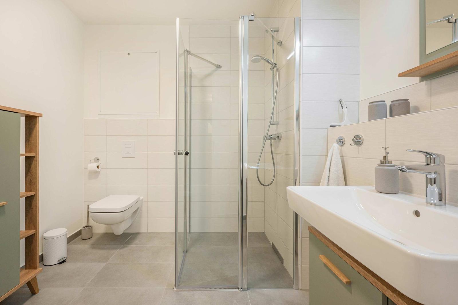 Badezimmer mit Dusche, Waschbecken und Toilette. Holzregal und graue Fliesen.