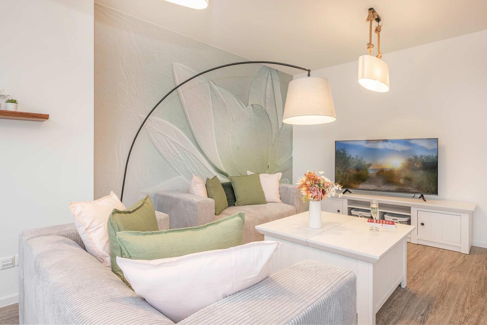 Wohnzimmer mit Sofa, Fernseher und Wandbild. Tisch mit Blumenvase und Gläsern.
