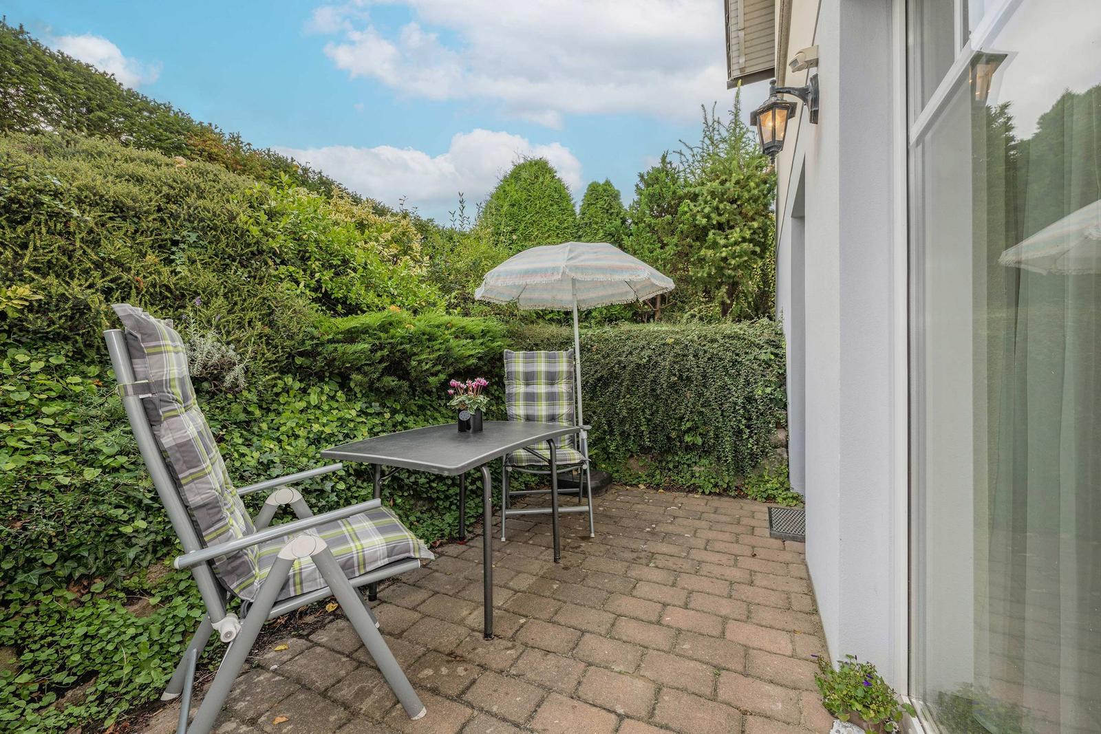 Terrasse mit Tisch, zwei Stühlen und Sonnenschirm. Hintergrund: grüne Hecke und blauer Himmel.