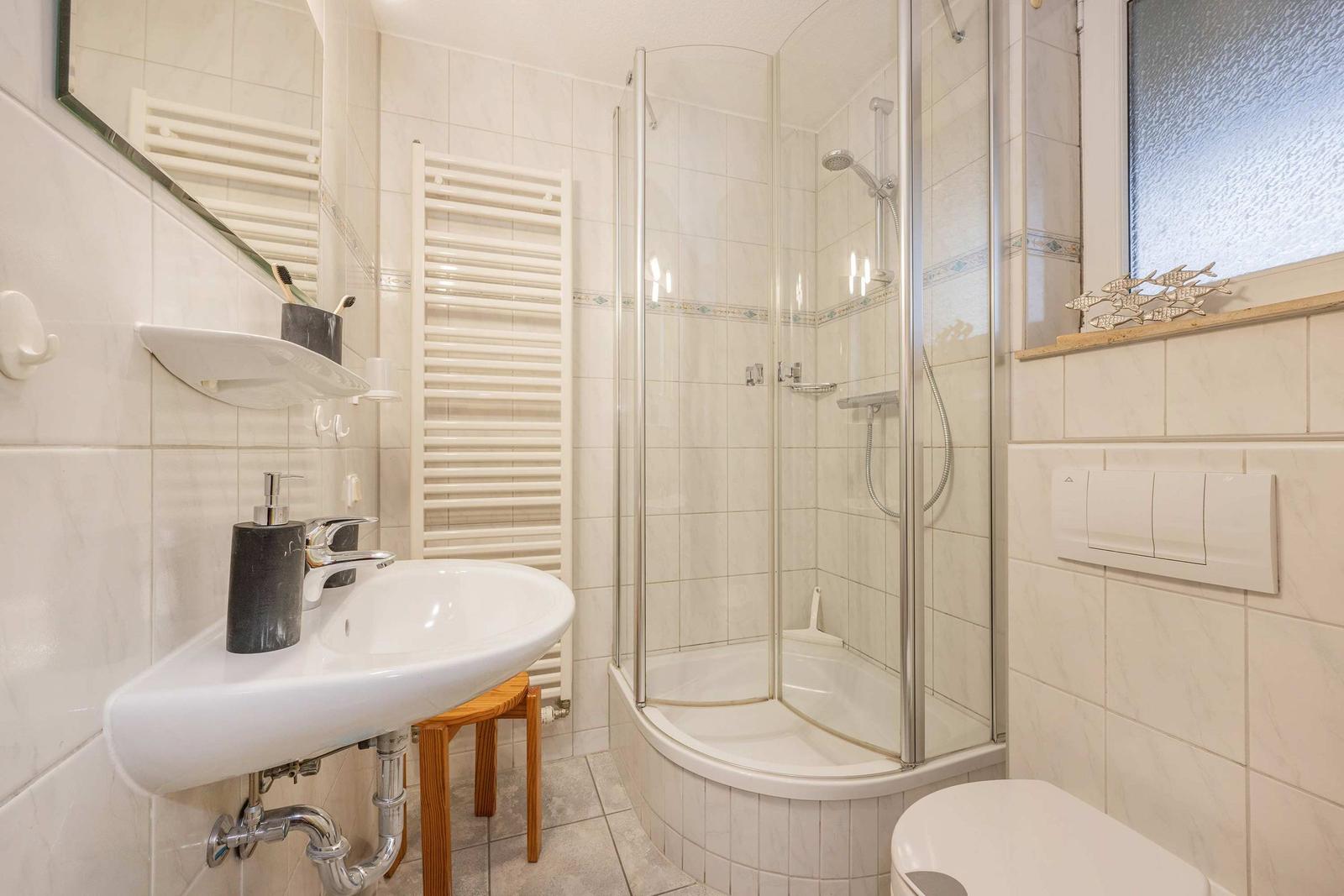 Badezimmer mit Dusche, Waschbecken und Toilette. Weiße Fliesen und Holzstuhl.