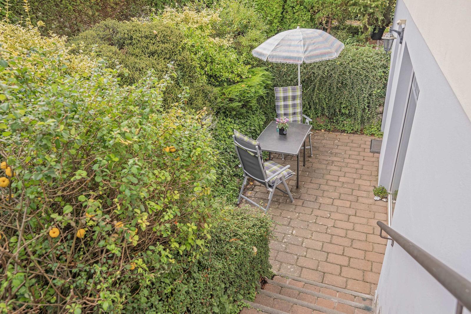 Terrasse mit Tisch, zwei Stühlen und Sonnenschirm zwischen grünen Hecken.